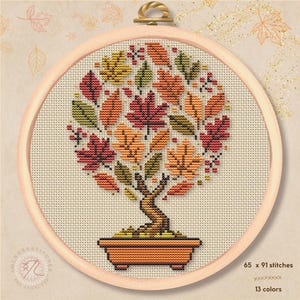 May include: A cross-stitch pattern in a round wooden hoop featuring a bonsai tree with autumn-colored leaves in shades of red, orange, yellow, and green. The tree is planted in a rectangular orange pot. The design measures 65 x 91 stitches.