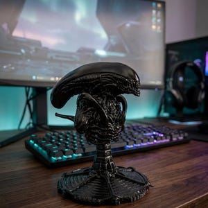 Pode incluir: Uma estatueta preta de Alien Xenomorph com detalhes intrincados. A estatueta está em uma mesa de madeira, com um monitor de computador e um teclado ao fundo. Um item colecionável.