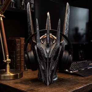 May include: A dark grey, metallic-finished helmet display stand featuring a tall, pointed, multi-spiked crown design with intricate, etched detailing. A pair of black over-ear headphones is resting securely on the sides of the helmet stand.