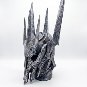 K&ouml;nnte beinhalten: Ein dunkler Metallhelm mit einer hohen, spitzen Krone und mehreren scharfen, vertikalen Dornen. Die Oberfl&auml;che ist mit komplizierten, ge&auml;tzten Rankenmustern verziert und weist ein verwittertes, abgenutztes Finish in Silber- und Anthrazitt&ouml;nen auf.