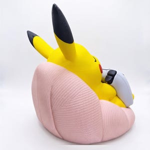 May include: A yellow Pikachu figure with black-tipped ears and red cheeks, sitting in a light pink, textured, shell-shaped chair. The figure is holding a white and black gaming controller in its hands, viewed from a side angle against a white background.
