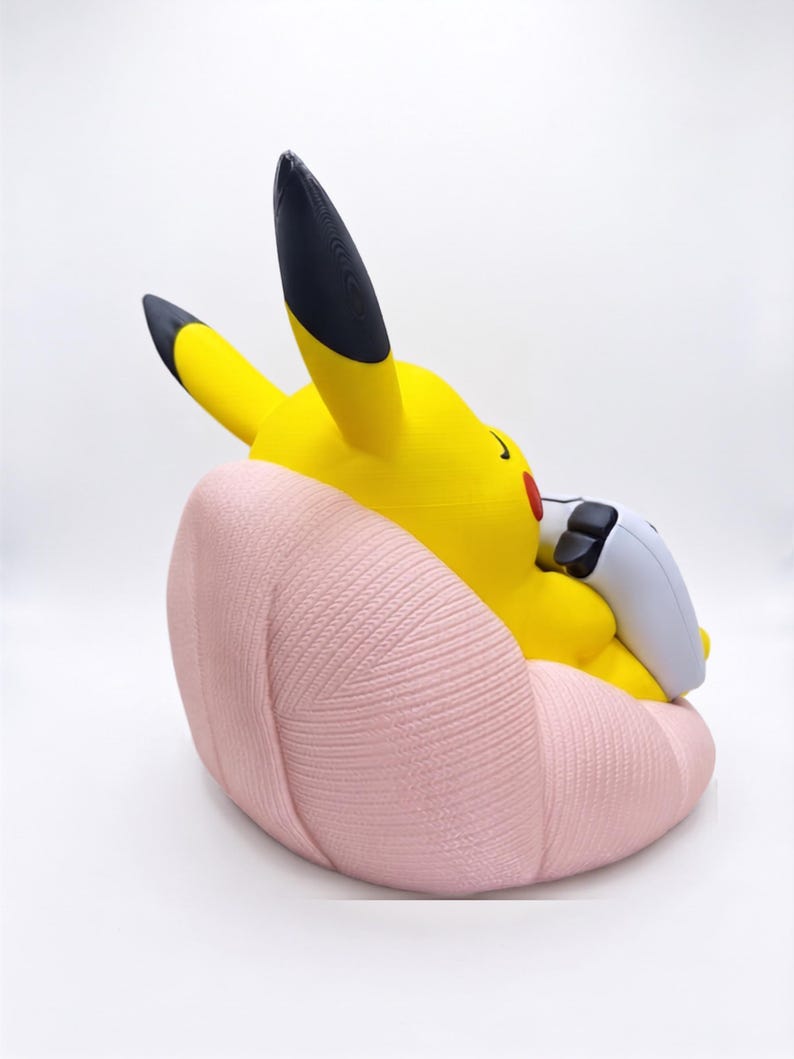 May include: A yellow Pikachu figure with black-tipped ears and red cheeks, sitting in a light pink, textured, shell-shaped chair. The figure is holding a white and black gaming controller in its hands, viewed from a side angle against a white background.