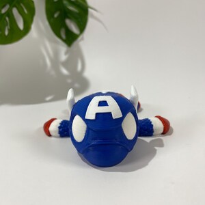 May include: A blue Captain America-themed figurine with white accents and the letter "A" on the front. The figurine has white horns and red and white striped arms and feet. The background is white with a green plant.