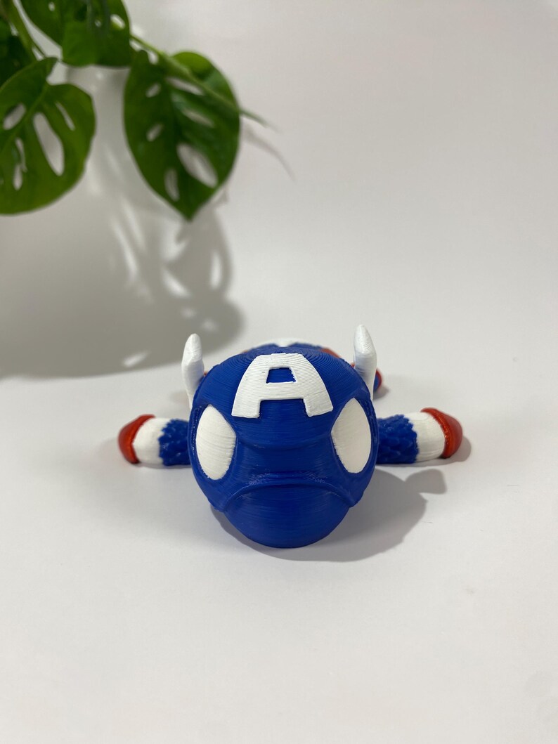 May include: A blue Captain America-themed figurine with white accents and the letter "A" on the front. The figurine has white horns and red and white striped arms and feet. The background is white with a green plant.
