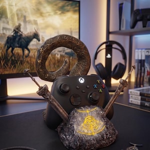 Op de afbeelding: Een zwarte Xbox-controller rust op een thematische standaard met een rotsbasis en twee zwaardachtige steunen. De standaard heeft een gouden embleem en een gebogen hoorndesign. Een monitor met een game-sc&egrave;ne is op de achtergrond te zien.