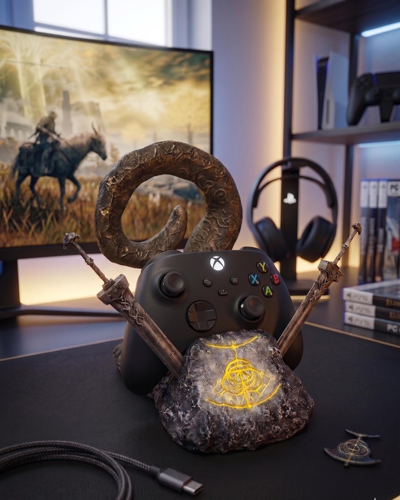 Op de afbeelding: Een zwarte Xbox-controller rust op een thematische standaard met een rotsbasis en twee zwaardachtige steunen. De standaard heeft een gouden embleem en een gebogen hoorndesign. Een monitor met een game-sc&egrave;ne is op de achtergrond te zien.