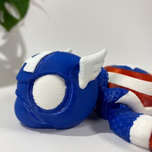 May include: A blue and white 3D-printed toy helmet with wing accents, reminiscent of a superhero. The helmet features a white "A" on the forehead and is partially visible, with a red, white, and blue striped section.