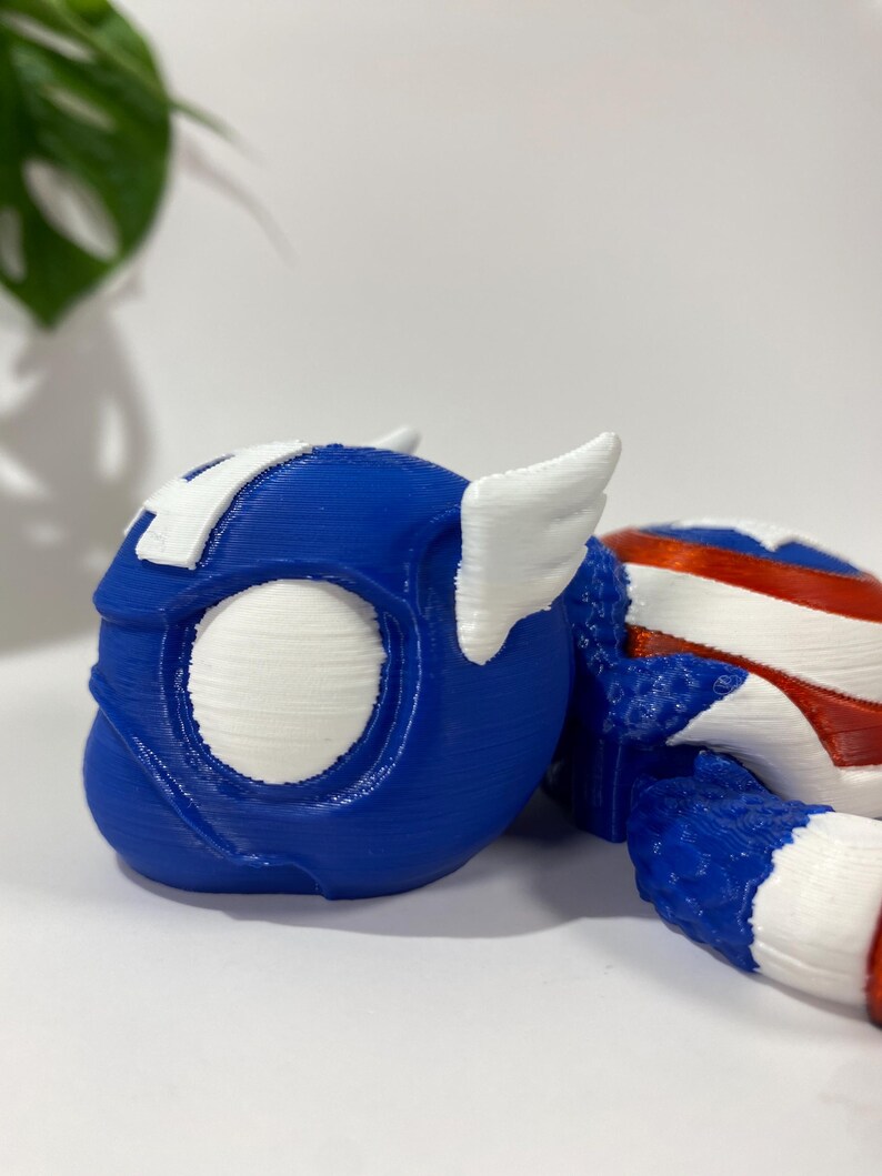May include: A blue and white 3D-printed toy helmet with wing accents, reminiscent of a superhero. The helmet features a white "A" on the forehead and is partially visible, with a red, white, and blue striped section.