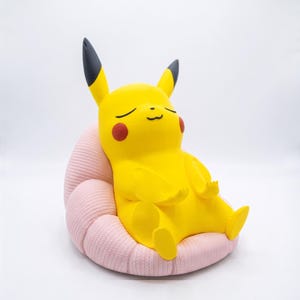 May include: A yellow Pikachu figure with black-tipped ears and red cheeks, sitting in a meditative pose with eyes closed. The figure is resting on a light pink, textured, rounded cushion that features a ribbed, fabric-like surface pattern.