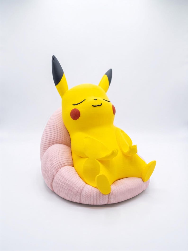 May include: A yellow Pikachu figure with black-tipped ears and red cheeks, sitting in a meditative pose with eyes closed. The figure is resting on a light pink, textured, rounded cushion that features a ribbed, fabric-like surface pattern.