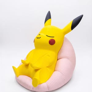 May include: A yellow Pikachu figure with black-tipped ears and red circular cheeks, sitting in a relaxed pose on a textured, light pink, rounded cushion. The figure has closed eyes and a small, simple mouth, appearing to be in a state of rest.