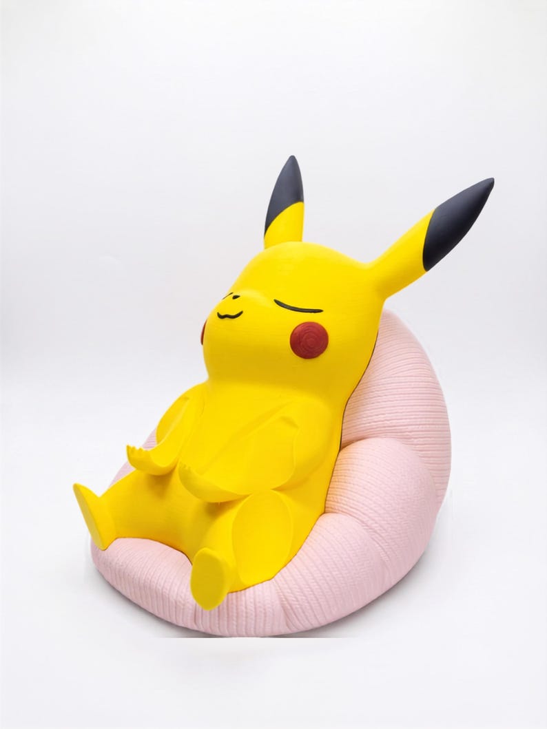 May include: A yellow Pikachu figure with black-tipped ears and red circular cheeks, sitting in a relaxed pose on a textured, light pink, rounded cushion. The figure has closed eyes and a small, simple mouth, appearing to be in a state of rest.