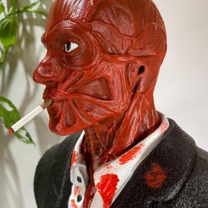 Pu&ograve; includere: Una scultura anatomica rossa di una testa umana, con dettagli muscolari esposti, che fuma una sigaretta. La figura indossa una giacca nera sopra una camicia bianca con schizzi rossi. La scultura ha un aspetto realistico e dettagliato.