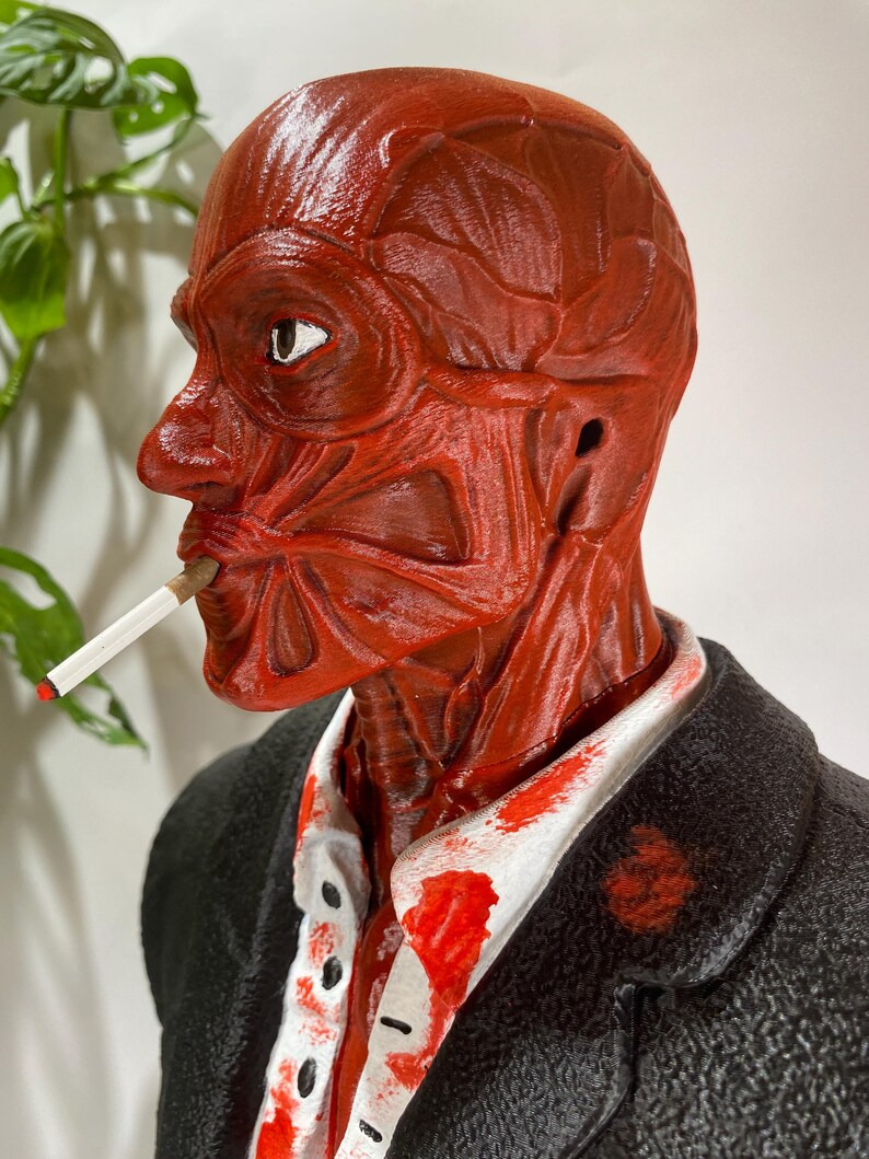 Pu&ograve; includere: Una scultura anatomica rossa di una testa umana, con dettagli muscolari esposti, che fuma una sigaretta. La figura indossa una giacca nera sopra una camicia bianca con schizzi rossi. La scultura ha un aspetto realistico e dettagliato.