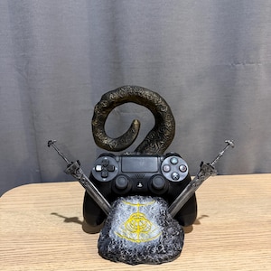 Op de afbeelding: Een zwarte PlayStation 4-controller rust op een stenen voet met een gouden embleem. Twee zilveren zwaarden kruisen elkaar achter de controller en een bronzen hoornvormig element rijst op.