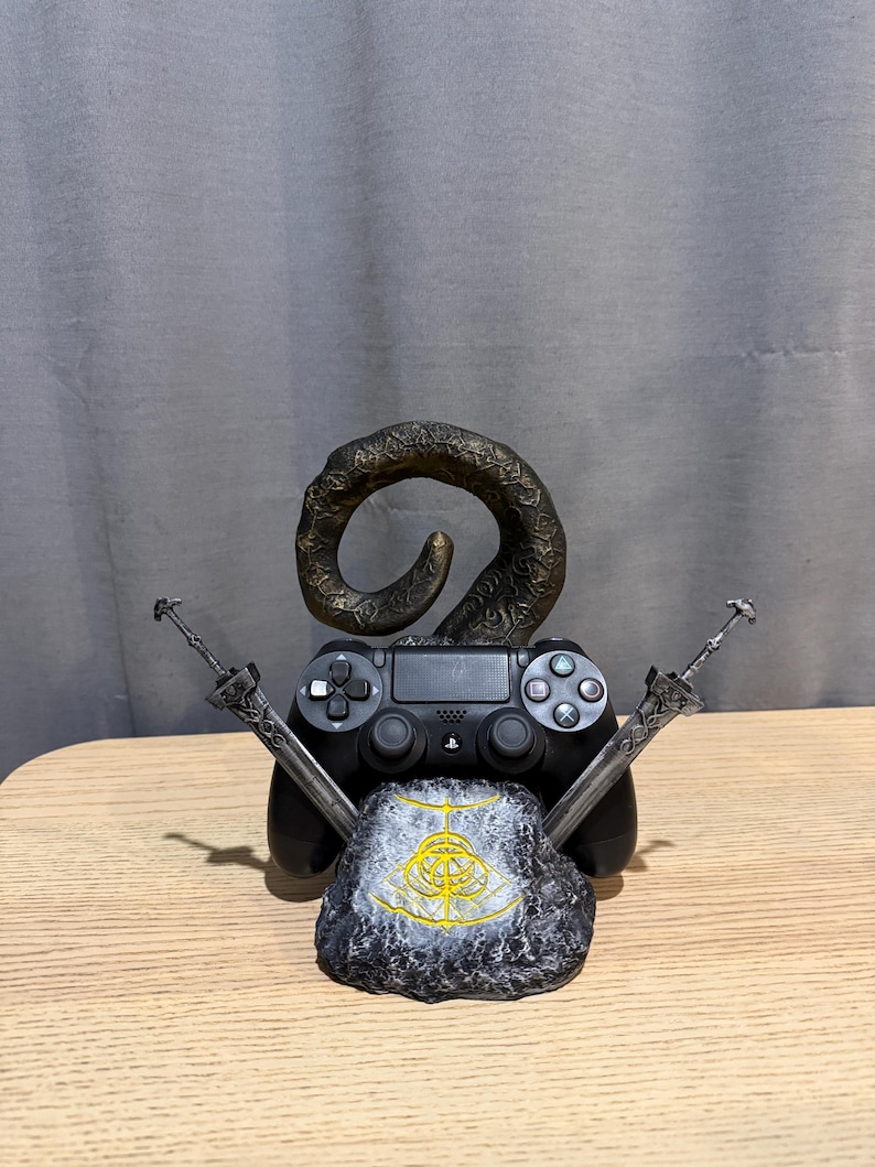 Op de afbeelding: Een zwarte PlayStation 4-controller rust op een stenen voet met een gouden embleem. Twee zilveren zwaarden kruisen elkaar achter de controller en een bronzen hoornvormig element rijst op.