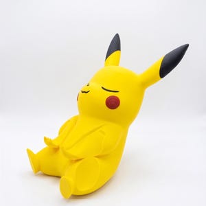 May include: A yellow Pikachu figurine in a seated, meditative pose with eyes closed. The character features long ears with black tips, round red cheek patches, and a small mouth. The figure is rendered in a smooth, matte finish with a minimalist design.