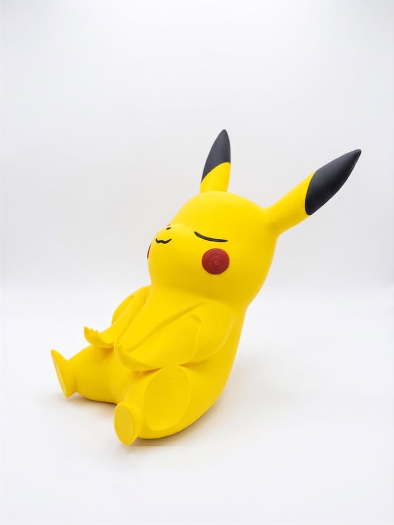 May include: A yellow Pikachu figurine in a seated, meditative pose with eyes closed. The character features long ears with black tips, round red cheek patches, and a small mouth. The figure is rendered in a smooth, matte finish with a minimalist design.