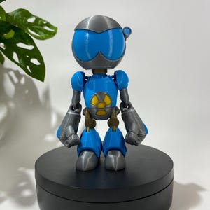 Puede incluir: Una figura de robot impresa en 3D de color azul y gris con un s&iacute;mbolo de radiaci&oacute;n amarillo en el pecho. El robot est&aacute; de pie sobre una base circular negra.