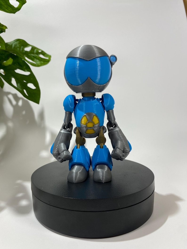 Puede incluir: Una figura de robot impresa en 3D de color azul y gris con un s&iacute;mbolo de radiaci&oacute;n amarillo en el pecho. El robot est&aacute; de pie sobre una base circular negra.