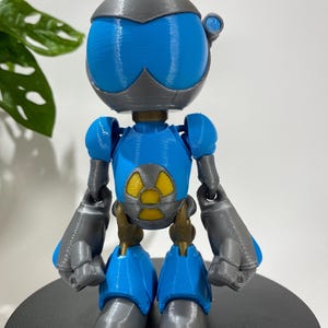 Puede incluir: Una figura de robot impresa en 3D azul y gris con un s&iacute;mbolo de radiaci&oacute;n amarillo en el pecho.