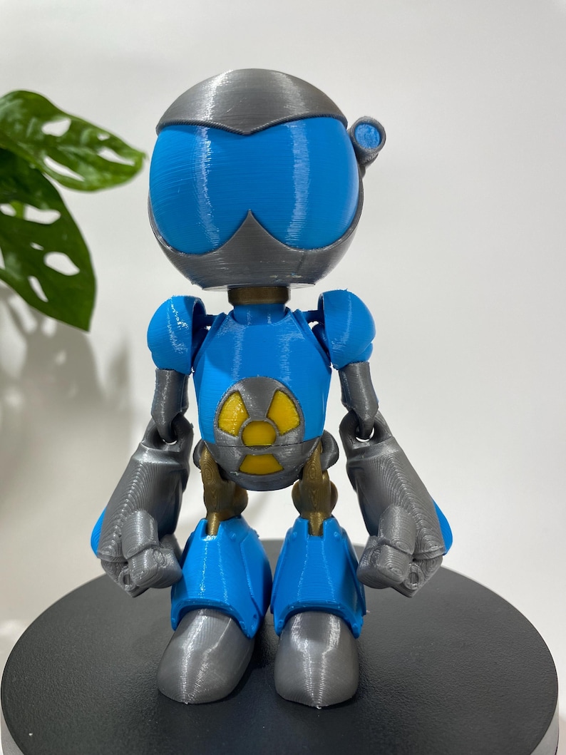 Puede incluir: Una figura de robot impresa en 3D azul y gris con un s&iacute;mbolo de radiaci&oacute;n amarillo en el pecho.