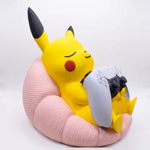 May include: A yellow Pikachu figure with black-tipped ears and red cheeks, sitting in a textured light pink beanbag chair. The figure is holding a white and black video game controller in its hands, with its eyes closed in a relaxed expression.