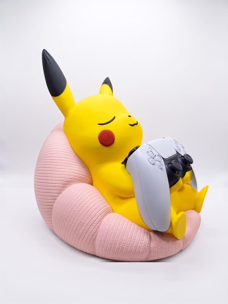 May include: A yellow Pikachu figure with black-tipped ears and red cheeks, sitting in a textured light pink beanbag chair. The figure is holding a white and black video game controller in its hands, with its eyes closed in a relaxed expression.