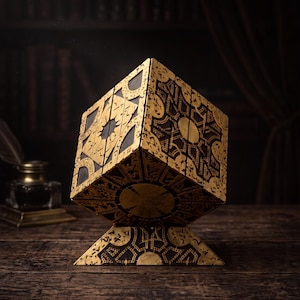 May include: A decorative puzzle box featuring a complex geometric design with gold-colored engravings on a dark brown base. The cube is balanced on a triangular stand, showcasing intricate patterns and circular motifs on each visible side panel.