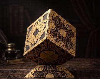 Puede incluir: Una caja de rompecabezas decorativa con un diseño geométrico complejo y grabados de color dorado sobre una base marrón oscuro. El cubo se equilibra sobre un soporte triangular, mostrando patrones intrincados en cada panel lateral.