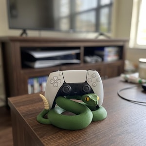 Könnte beinhalten: Ein grüner, schlangenförmiger Controller-Ständer mit einer cremefarbenen Rassel am Schwanz und gelben Augen, der einen weißen und schwarzen kabellosen Game-Controller hält. Die Schlange ist zu einer runden Basis gewunden, um den Controller zu stützen.