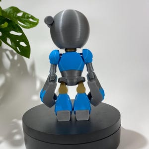 Puede incluir: Una figura de robot impresa en 3D con una cabeza gris, un cuerpo azul y brazos y piernas grises. El robot est&aacute; de pie sobre una base circular negra.