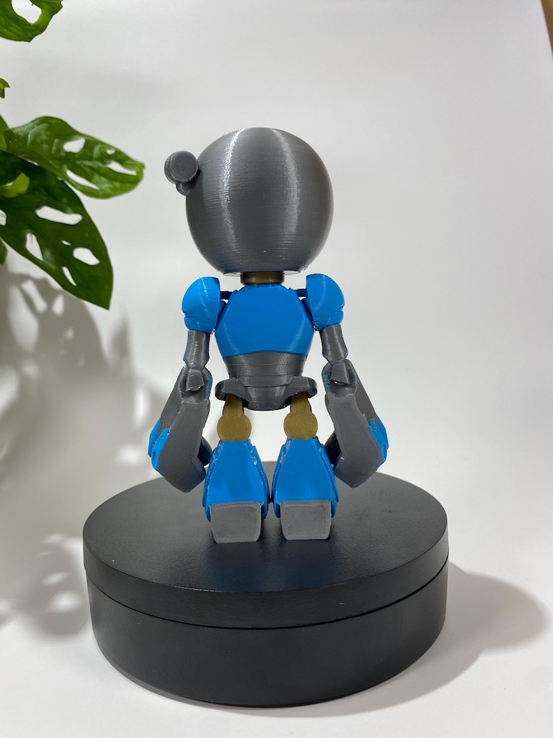 Puede incluir: Una figura de robot impresa en 3D con una cabeza gris, un cuerpo azul y brazos y piernas grises. El robot est&aacute; de pie sobre una base circular negra.