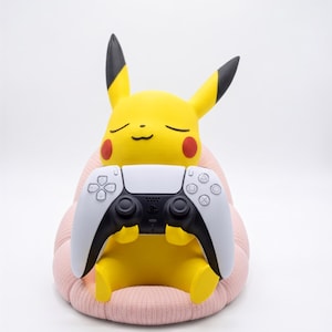 May include: A yellow Pikachu figure with black-tipped ears and red cheeks, sitting on a textured light pink cushion. The figure is holding a white and black wireless video game controller with a central touchpad and various circular buttons.