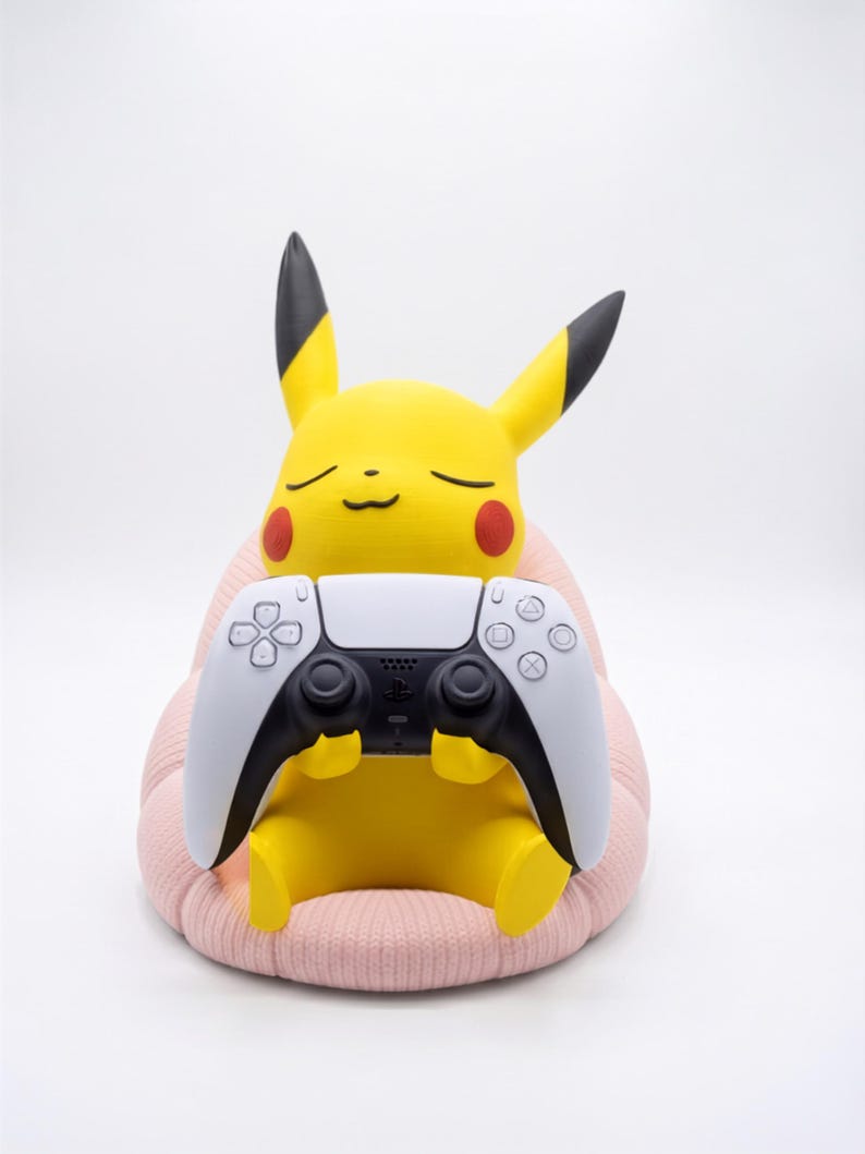 May include: A yellow Pikachu figure with black-tipped ears and red cheeks, sitting on a textured light pink cushion. The figure is holding a white and black wireless video game controller with a central touchpad and various circular buttons.