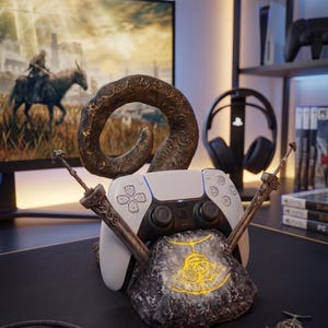 May include: A white and black PlayStation 5 controller rests on a stone base with a glowing yellow design. Two decorative swords and a bronze horn accent the controller. The setup is on a black desk, with a monitor in the background.