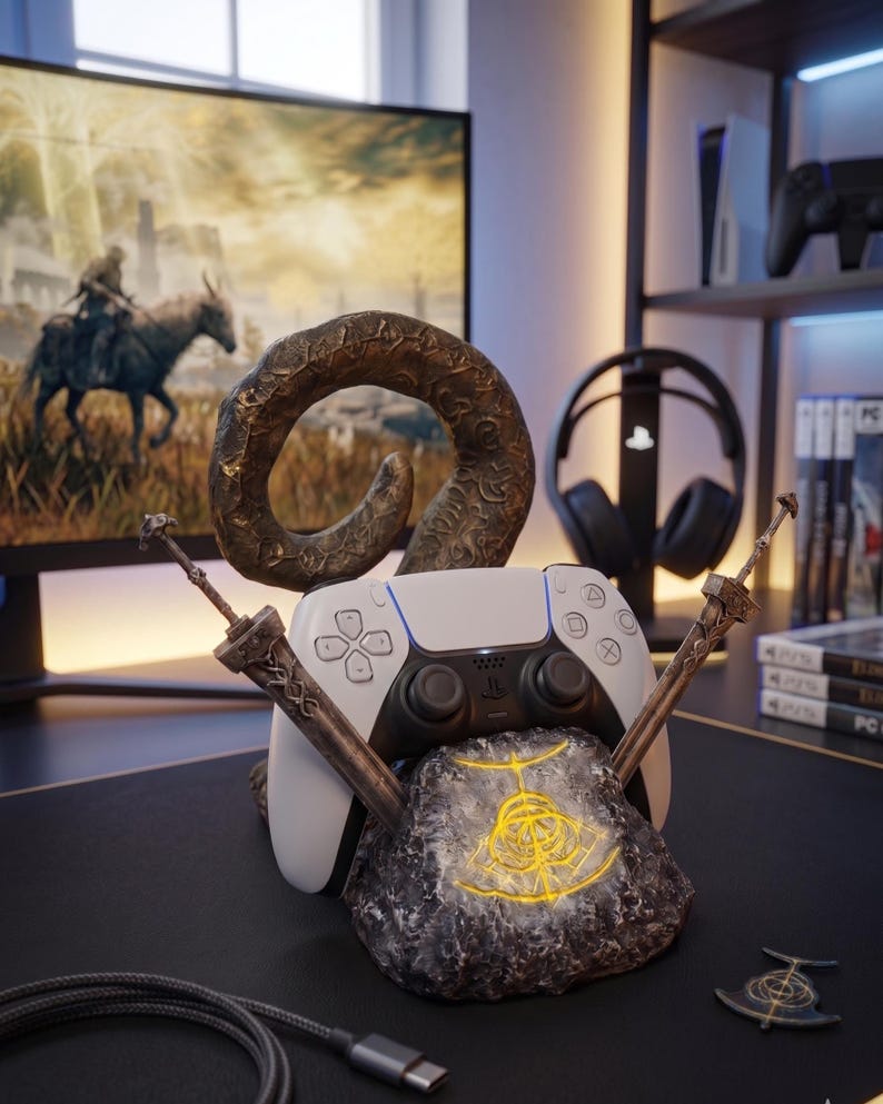 Op de afbeelding: Een witte en zwarte PlayStation 5-controller rust op een stenen basis met een gloeiend geel ontwerp. Twee decoratieve zwaarden en een bronzen hoorn accentueren de controller. De opstelling staat op een zwart bureau, met een monitor op de achtergrond.