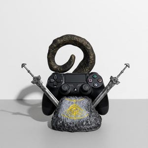 May include: A black PlayStation 4 controller rests on a stone base with a gold emblem. Two silver swords are crossed behind the controller. A dark, curved horn shape sits above the controller, completing the gaming-themed display.
