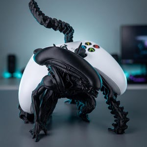 Könnte beinhalten: Eine schwarze Xenomorph-Figur, die als Controller-Ständer für einen weißen kabellosen Xbox-Controller dient. Die Kreatur befindet sich in einer hockenden Position mit ihrem langen, nach oben gebogenen Schwanz, der den Controller stützt.