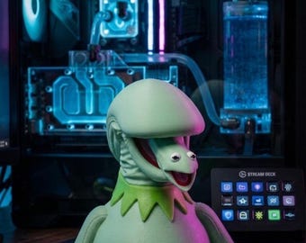 May include: A light green Kermit the Frog figurine with an open mouth, set against a computer with blue liquid cooling. A Stream Deck control panel is visible. A black mouse is on the desk.