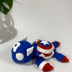 May include: A 3D-printed Captain America figurine, featuring a blue helmet with a white "A" and wings, and a body with a shield design. The figurine is primarily blue, red, and white.