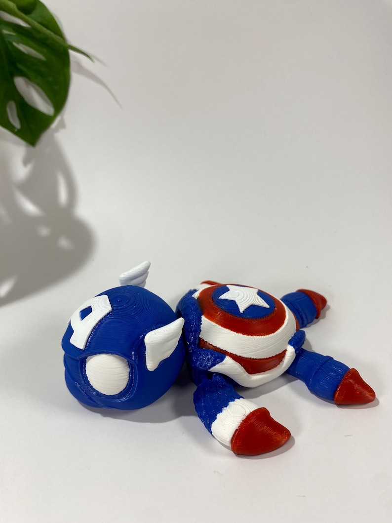 May include: A 3D-printed Captain America figurine, featuring a blue helmet with a white "A" and wings, and a body with a shield design. The figurine is primarily blue, red, and white.