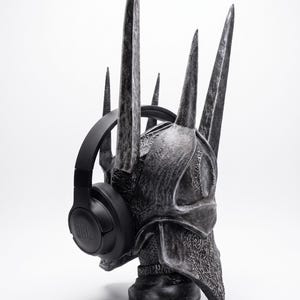 K&ouml;nnte beinhalten: Ein dunkelgrauer, texturierter Helm mit mehreren hohen, spitzen Zacken, einer verwitterten Oberfl&auml;che und komplexen Details. Ein mattschwarzer JBL-Over-Ear-Kopfh&ouml;rer ist &uuml;ber den Seiten der Helmstruktur angebracht.