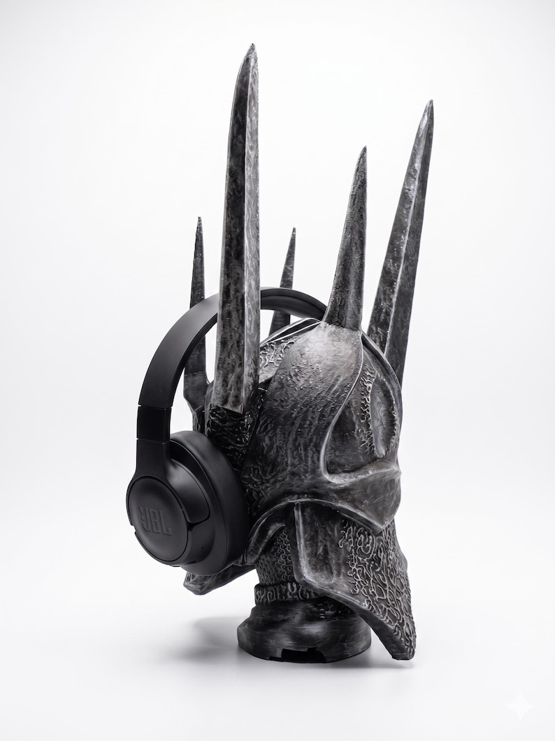 K&ouml;nnte beinhalten: Ein dunkelgrauer, texturierter Helm mit mehreren hohen, spitzen Zacken, einer verwitterten Oberfl&auml;che und komplexen Details. Ein mattschwarzer JBL-Over-Ear-Kopfh&ouml;rer ist &uuml;ber den Seiten der Helmstruktur angebracht.