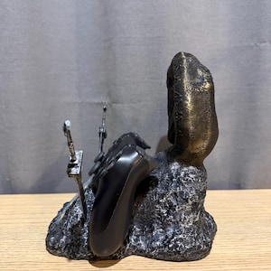 Op de afbeelding: Een zwarte en bronzen gamecontrollerhouder. De houder heeft een getextureerde, rotsachtige basis met een bronzen pilaar aan de rechterkant. Twee zilverkleurige zwaardachtige objecten bevinden zich aan de linkerkant. Een zwarte gamecontroller rust op de houder.