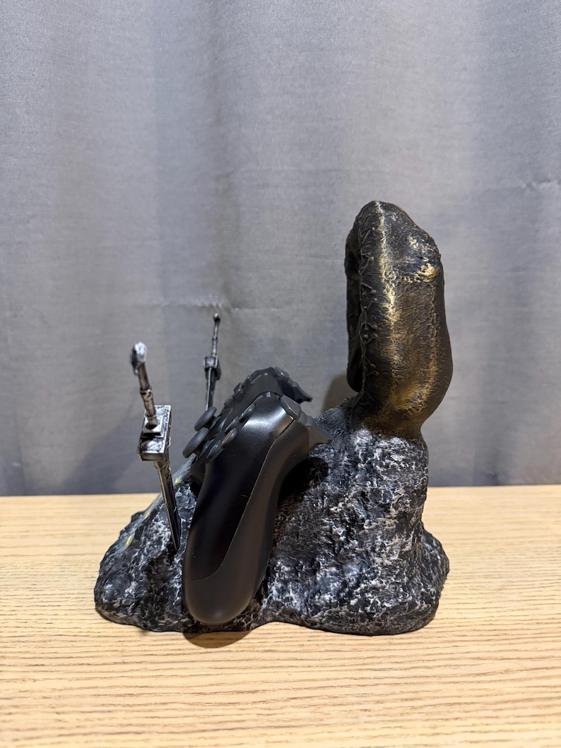 Op de afbeelding: Een zwarte en bronzen gamecontrollerhouder. De houder heeft een getextureerde, rotsachtige basis met een bronzen pilaar aan de rechterkant. Twee zilverkleurige zwaardachtige objecten bevinden zich aan de linkerkant. Een zwarte gamecontroller rust op de houder.