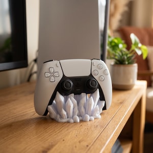 Könnte beinhalten: Ein weiß-schwarzer Videospiel-Controller ruht auf einem weißen, kristallförmigen Ständer. Der Controller verfügt über ein zentrales Touchpad, zwei Analogsticks und Standard-Aktionstasten. Der Ständer hat eine zerklüftete Textur.