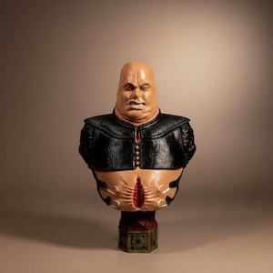 May include: A grotesque bust sculpture of a bald, fleshy figure with a stern expression. The figure wears a black leather-like jacket with gold buttons. The sculpture is mounted on a decorative, geometric base.
