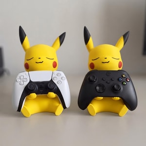 May include: Two yellow Pikachu figures with pointed ears and red cheeks, each sitting and holding a video game controller. One figure holds a white and black controller, while the other holds a solid black controller with colored buttons.