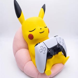 May include: A yellow Pikachu figure with black-tipped ears and red cheeks, sitting in a textured light pink chair. The figure holds a white and black wireless game controller in its hands, with its eyes closed in a relaxed, smiling expression.